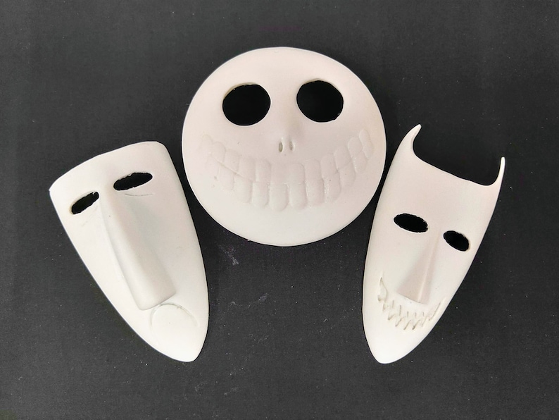 May include: Three white ceramic masks with different facial expressions. The masks are all in the shape of a human face, but with exaggerated features. The first mask has a wide smile with teeth, the second mask has a neutral expression, and the third mask has a frown.