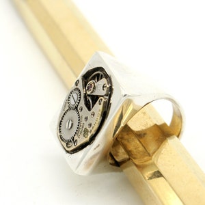 May include: A silver ring with a square face featuring a visible watch mechanism. The ring is set on a gold surface.