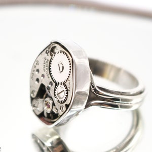 May include: A silver ring with a watch face embedded in the top. The watch face is exposed, showing the inner workings of the watch. The ring is designed to be worn on the finger.