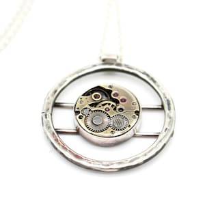 Puede incluir: Un collar de plata con un colgante redondo que presenta un marco circular con un pequeño mecanismo de relojería intrincado en su interior. El mecanismo de relojería es visible a través del marco y tiene pequeñas joyas rojas.