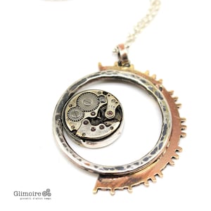Puede incluir: Un collar de cadena de plata con un colgante circular que presenta un mecanismo de reloj vintage. El colgante está hecho de plata y cobre, con una textura martillada. El mecanismo del reloj es visible a través de una ventana de vidrio transparente.