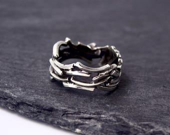 May include: A silver ring with a detailed, interwoven design. The ring features an intricate pattern of overlapping bars and curved elements, creating a unique, artistic appearance. The ring is shown on a dark grey, textured surface.