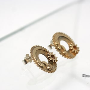 Peut inclure: Une paire de boucles d'oreilles dorées avec un motif d'engrenage. Chaque boucle d'oreille présente un engrenage circulaire avec un plus petit engrenage qui y est attaché. Les engrenages sont finement détaillés et ont une finition polie.