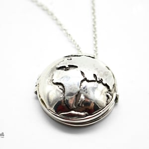 May include: A silver-colored globe pendant necklace. The pendant is a detailed representation of the Earth, with continents and landmasses visible. The necklace chain is also silver. The pendant is round and has a textured surface.