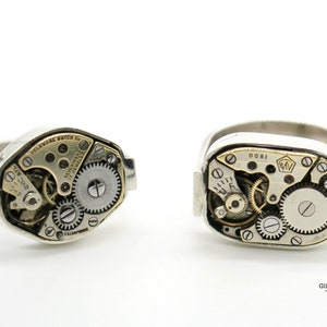 Puede incluir: Dos anillos de plata con mecanismos de reloj expuestos. El anillo de la izquierda tiene el texto "DELAWARE WATCH CO. 17 JEWELS" en la cara. El anillo de la derecha tiene el texto "1800" en la cara.