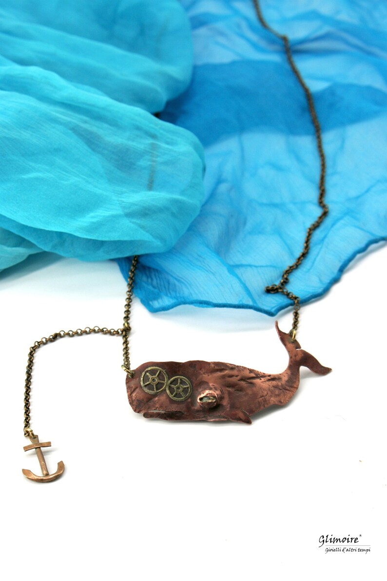 May include: A copper whale pendant necklace with brass gears and an anchor charm. The necklace is displayed on a blue fabric background.