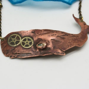May include: A copper whale pendant necklace with brass gears for eyes. The whale is a stylized silhouette with a textured surface. The necklace is on a chain.