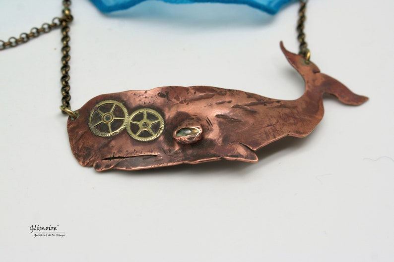 May include: A copper whale pendant necklace with brass gears for eyes. The whale is a stylized silhouette with a textured surface. The necklace is on a chain.