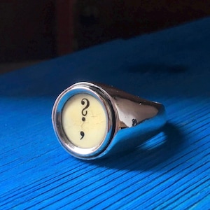 May include: A polished silver ring with a circular face displaying a question mark and comma on a cream-coloured background. The ring reflects light and rests on a blue surface.