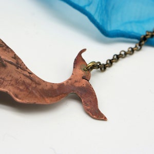 May include: A copper whale tail pendant necklace with a brass chain. The whale tail is textured and has a rustic look. The chain is attached to the top of the tail.