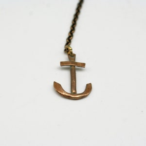 May include: A copper anchor pendant hangs from a chain. The anchor is a simple design with a cross at the top and a curved bottom.