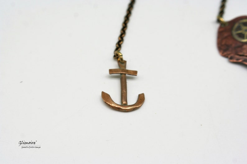 May include: A copper anchor pendant hangs from a chain. The anchor is a simple design with a cross at the top and a curved bottom.