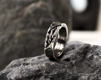May include: A silver ring with a braided rope design. The ring is set on a dark grey rock, showcasing its intricate details and metallic sheen. The ring's design features a textured surface.