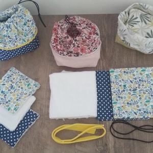 Op de afbeelding: Een set handgemaakte stoffen artikelen, waaronder drie trekkoordtassen, drie stoffen vierkanten, een witte washandje, een gele elastiek en een zwart haarbandje. De tassen zijn versierd met bloemenpatronen en de vierkanten zijn gemaakt van een blauw-wit geblokte stof met een bloemenpatroon.