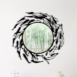 May include: A round mirror with a black metal frame featuring a design of stylized fish. The mirror is hanging on a white wall above a white dresser with three drawers.