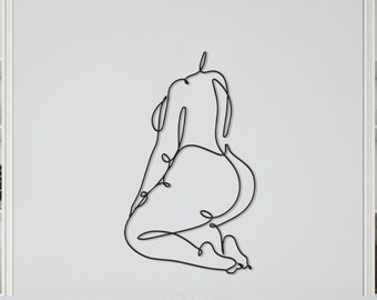 May include: A black metal wall art piece depicting a continuous line drawing of a person in a kneeling pose. The artwork is mounted on a white wall above a white sofa, creating a minimalist aesthetic. The room features white shelves with books and decorative items.