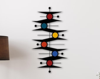 May include: A black metal wall art piece with a mid-century modern design. The artwork features a vertical arrangement of black geometric shapes, red, blue, and yellow circles, and thin horizontal lines. The piece is mounted on a white wall.