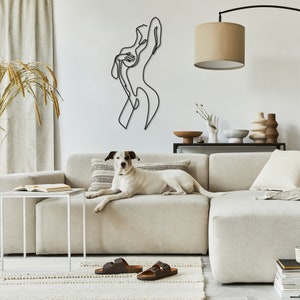 May include: A white dog is lying on a light beige sectional sofa. The sofa is in front of a white wall with a black metal line art sculpture of a woman. The sculpture is on the wall above a white coffee table.
