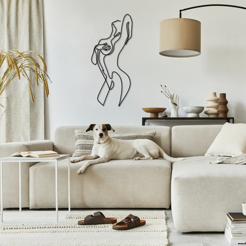 May include: A white dog is lying on a light beige sectional sofa. The sofa is in front of a white wall with a black metal line art sculpture of a woman. The sculpture is on the wall above a white coffee table.