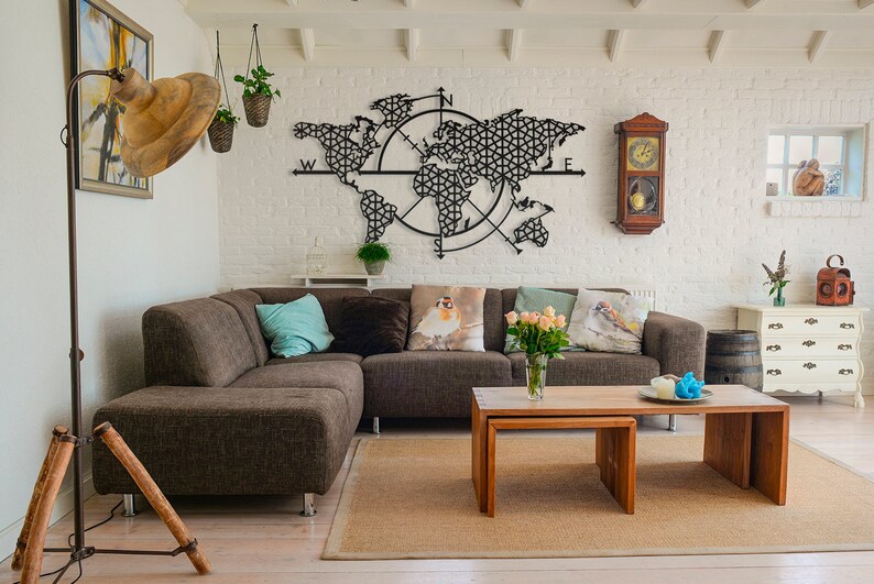 May include: A brown sectional sofa with pillows sits in front of a black metal world map wall decor with a compass design. The room has white brick walls, a wooden floor, and a wooden coffee table with a vase of pink roses.
