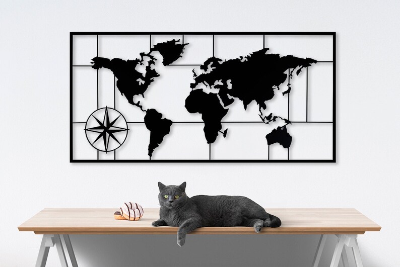 May include: Black metal wall art featuring a world map with a compass in the lower left corner. The map is outlined with a grid and the continents are cut out. A gray cat is lying on a wooden table in the foreground.