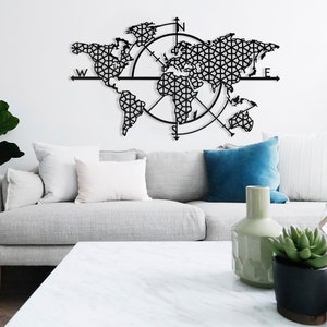 May include: A black metal world map wall decor with a compass design. The map is made of intricate geometric patterns and is surrounded by a compass rose with the letters N, S, E, and W. The map is hanging on a white wall above a gray couch with white pillows and a blue pillow.