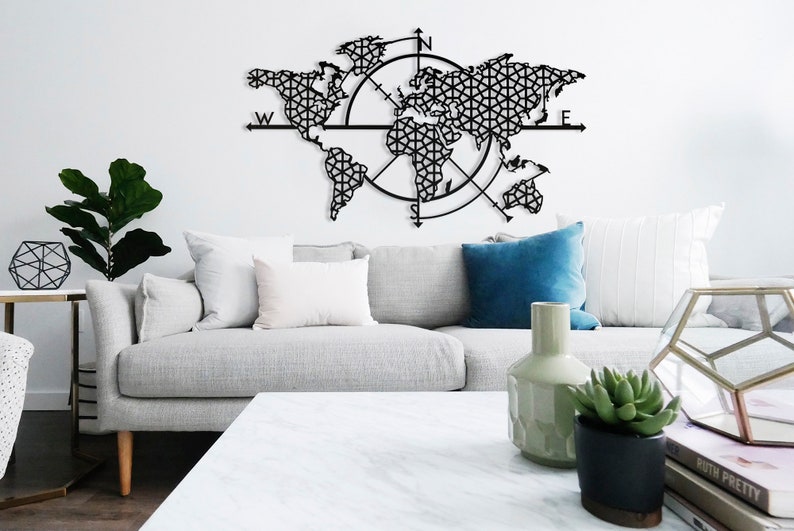May include: A black metal world map wall decor with a compass design. The map is made of intricate geometric patterns and is surrounded by a compass rose with the letters N, S, E, and W. The map is hanging on a white wall above a gray couch with white pillows and a blue pillow.