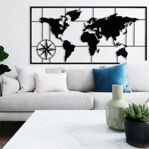 May include: A black metal world map wall decor with a compass design. The map is framed in a black metal frame and is mounted on a white wall. The map is displayed above a gray couch with white pillows and a blue pillow. There is a coffee table in front of the couch with a vase and a plant on it.