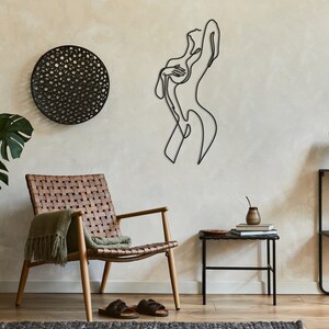 May include: A black metal wall sculpture of a woman in a single line art style. The sculpture is mounted on a beige wall with a woven circular wall hanging to the left. A brown leather chair with a green throw blanket is in the foreground.
