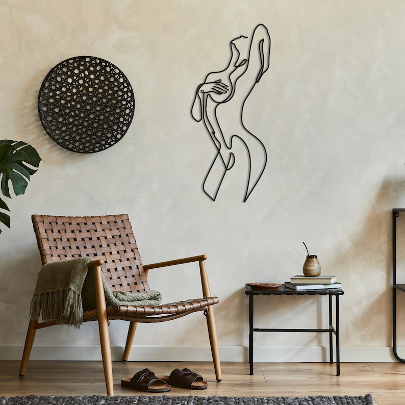 May include: A black metal wall sculpture of a woman in a single line art style. The sculpture is mounted on a beige wall with a woven circular wall hanging to the left. A brown leather chair with a green throw blanket is in the foreground.