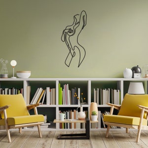 May include: A black metal wall sculpture of a stylized female figure in a single line design. The sculpture is mounted on a light green wall above a white bookshelf filled with books.