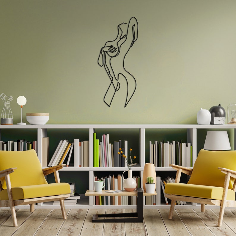 May include: A black metal wall sculpture of a stylized female figure in a single line design. The sculpture is mounted on a light green wall above a white bookshelf filled with books.