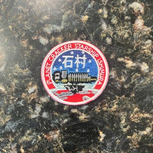 May include: A red, white, and blue embroidered patch with the text "Planet Cracker Starship Ishumura" and the Chinese character "石村" in the center. The patch features a stylized image of a spaceship with a rocket engine.