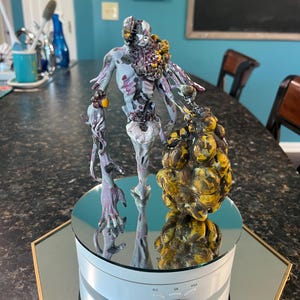 Może przedstawiać: Szczegółowa, ręcznie malowana figurka potwornego stworzenia z wieloma kończynami i mięsistym, bulwiastym wyrostkiem. Stworzenie jest pomalowane w odcieniach szarego, fioletowego i żółtego i jest wystawione na obracającej się platformie z lustrzaną powierzchnią.