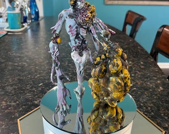 May include: A hand-painted figurine of a humanoid creature with a pale, mottled body, featuring a large, textured, yellow and brown growth on its left side and shoulder, standing on a white, circular, motorised rotating display stand.