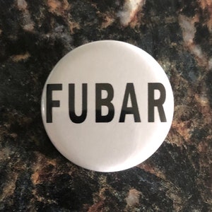 Puede incluir: Un botón blanco con la palabra "FUBAR" impresa en letras negras en negrita. El botón es redondo y parece ser de metal o plástico. El fondo es una superficie oscura y texturizada.