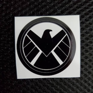 Può includere: Adesivo bianco e nero con il logo della fittizia organizzazione S.H.I.E.L.D. dell'Universo Cinematografico Marvel. Il logo è un cerchio con un'aquila stilizzata all'interno e una stella al centro.