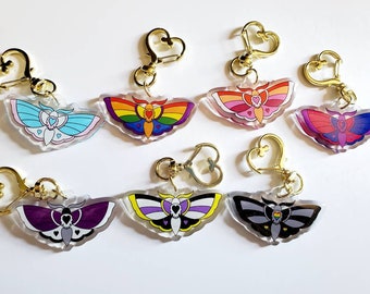 May include: Seven colorful moth keychains with gold heart-shaped rings. Each keychain features a different color moth with a heart design on its wings. The moths are blue, rainbow, pink, red, black, white, and purple.