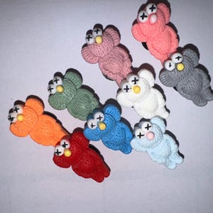 May include: A set of nine plush toys in various colors, each featuring a three-eyed monster design with an X for eyes. The toys are made of soft, fuzzy material and have a slightly distressed look.