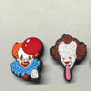 May include: Two colorful rubber charms featuring the character Pennywise from the movie "It". One charm shows Pennywise with a red balloon, the other shows Pennywise with his tongue sticking out.