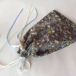 May include: A small gray fabric bag with a floral pattern in shades of yellow, white, and blue. The bag has white and blue ribbon ties.