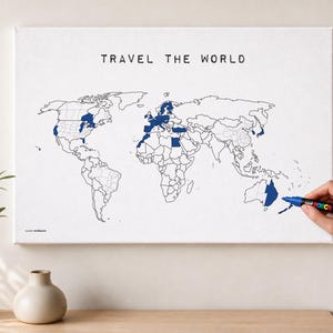 Puede incluir: Un mapa de pared en lienzo blanco con el texto TRAVEL THE WORLD en letras negras. El mapa muestra contornos negros de continentes y países, con varias regiones rellenas de un color azul oscuro. Un marcador negro está coloreando Australia.