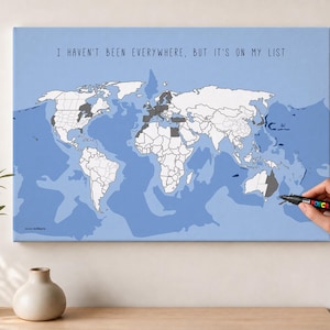 Könnte beinhalten: Eine rechteckige Wandleinwand mit einer Weltkarte auf hellblauem Ozeanhintergrund. Die Kontinente sind weiß umrandet, einige Regionen sind dunkelgrau schattiert. Der Text "I HAVEN'T BEEN EVERYWHERE, BUT IT'S ON MY LIST" ist oben in Schwarz gedruckt.