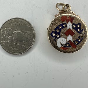May include: A gold-colored, round pendant with a patriotic design featuring red and white stripes and blue stars. The pendant is next to a North Dakota quarter.