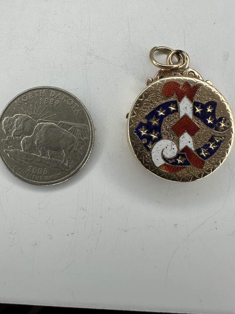 May include: A gold-colored, round pendant with a patriotic design featuring red and white stripes and blue stars. The pendant is next to a North Dakota quarter.