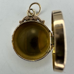 May include: Open gold-colored locket pendant with a hinged design. The locket is round with a decorative top and a loop for a chain. The interior is visible, revealing a hollow space. The locket is open, showing the inside.