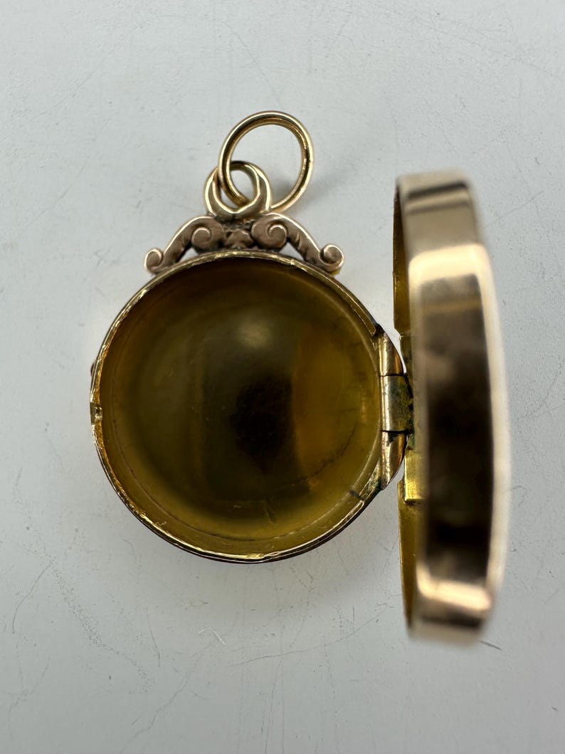 May include: Open gold-colored locket pendant with a hinged design. The locket is round with a decorative top and a loop for a chain. The interior is visible, revealing a hollow space. The locket is open, showing the inside.