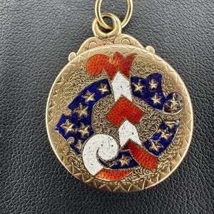 May include: A gold-colored, round pendant with a detailed design. The pendant features a central design in red, white, and blue, with a pattern of stars. The pendant has a loop at the top for attaching to a chain.