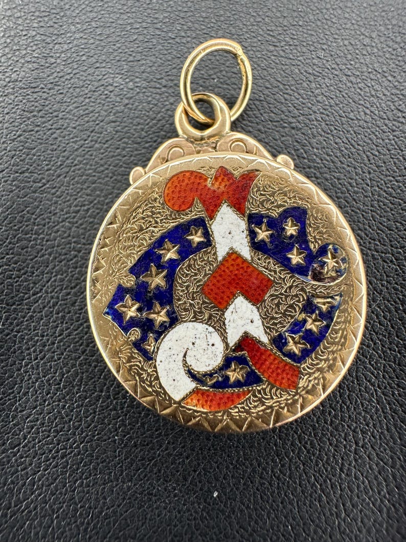 May include: A gold-colored, round pendant with a detailed design. The pendant features a central design in red, white, and blue, with a pattern of stars. The pendant has a loop at the top for attaching to a chain.