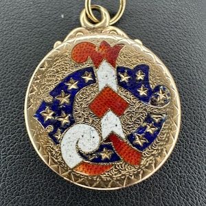May include: A gold-toned pendant with a detailed design. The pendant features a central design in red, white, and blue, with a pattern of stars. The pendant has a loop at the top for attaching to a chain.